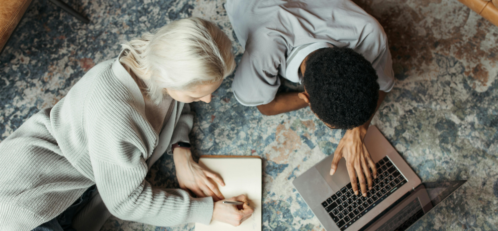 Kaksi opiskelijaa rennosti lattialla tekemässä työtä yhdessä, toinen tietokoneella, toisella kynä ja paperia